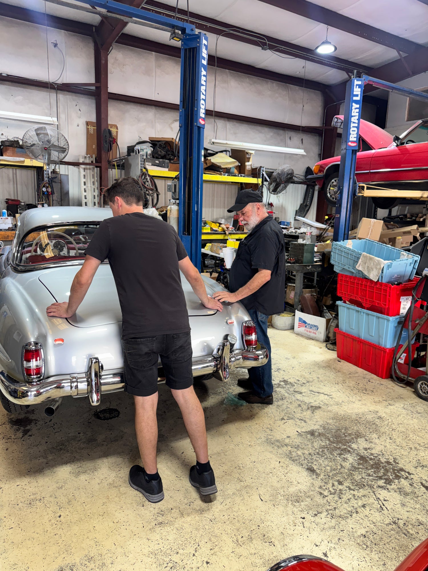 Two mechanics working on a classic vehicle under a lift in a garage.