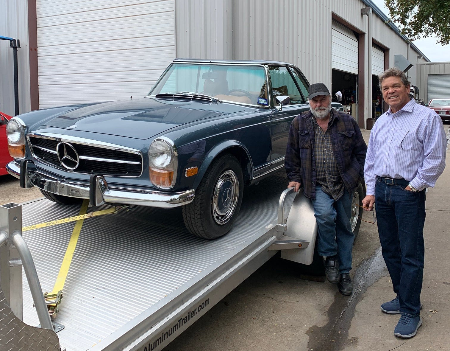 Two men standing next to a vintage Mercedes Benz car on a car trailer in front of a workshop