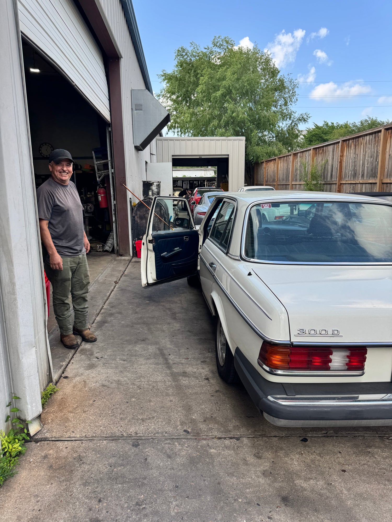 Mechanic smiling outside a mechanic shop next to a white Mercedes 300 D