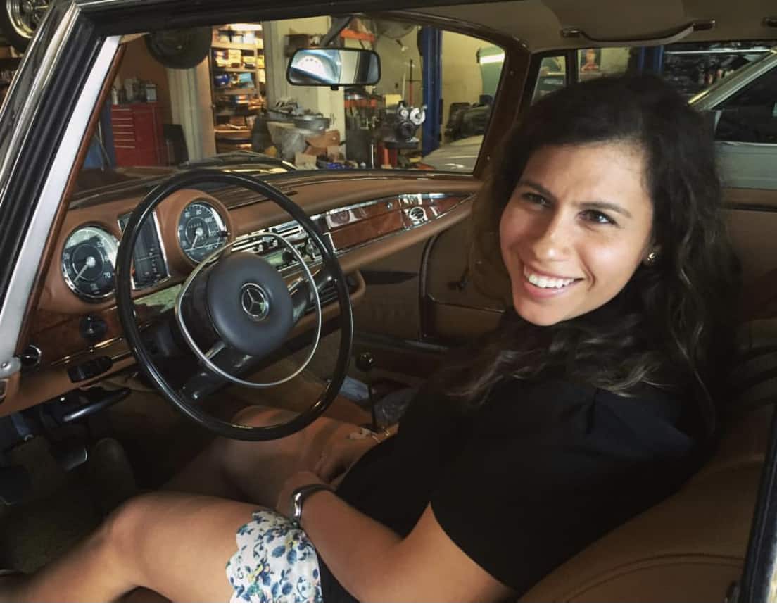 Woman sitting inside a vintage Mercedes-Benz car smiling at the camera.