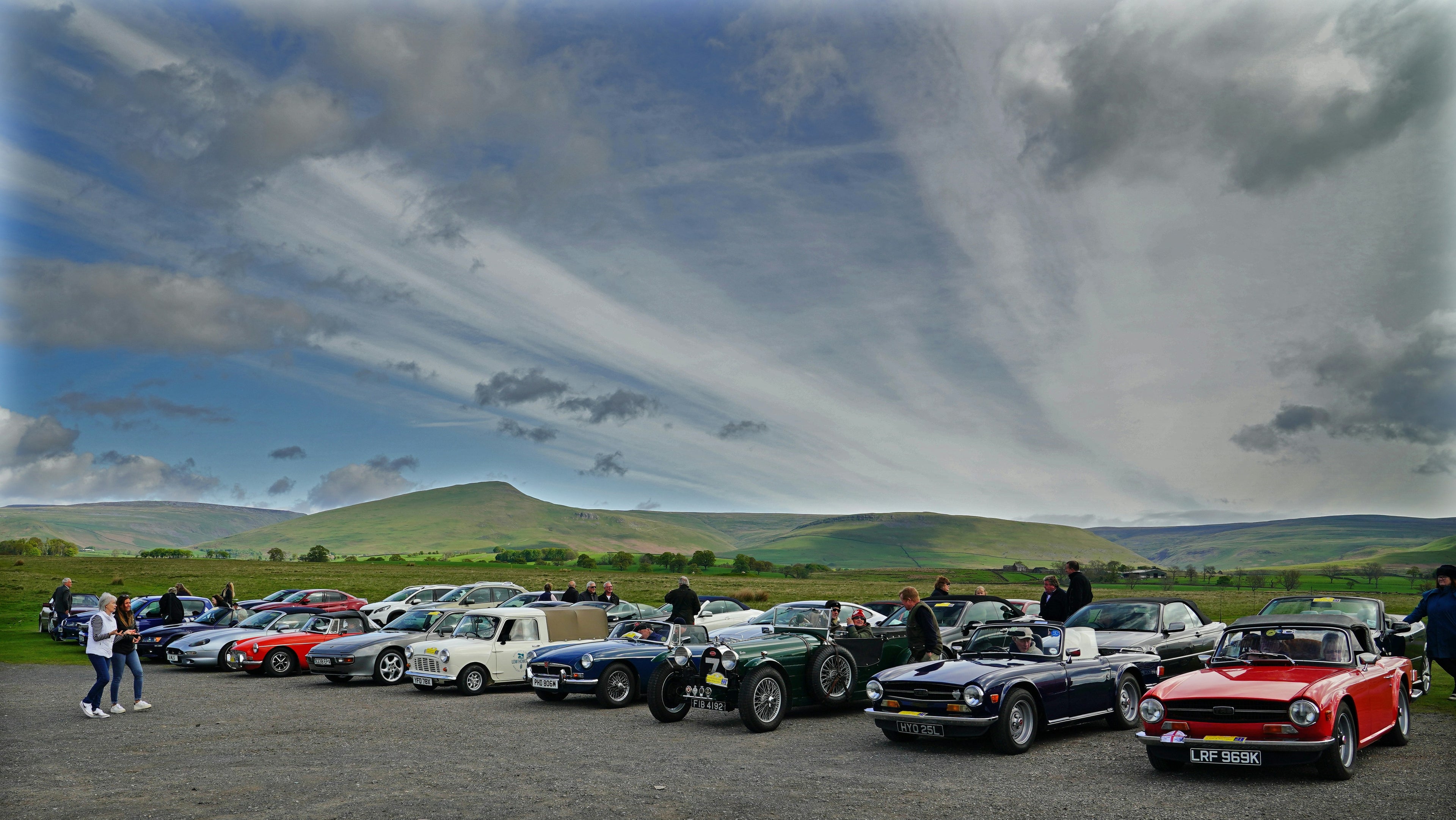 Collection of classic cars parked on a gravel surface with a scenic background